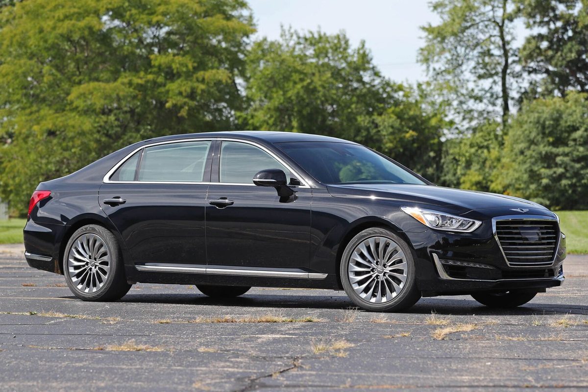 A black first-generation Genesis G90 sedan with chrome trim and multi-spoke wheels, parked on cracked pavement with trees behind