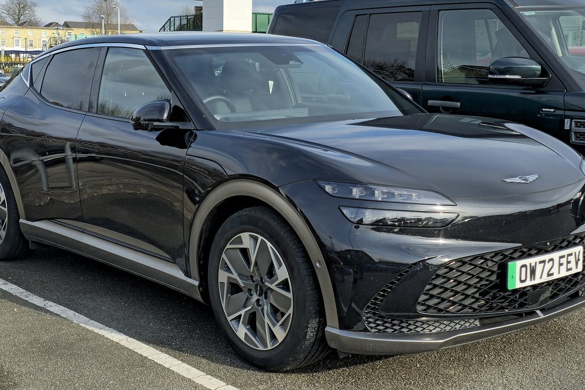 A black Genesis GV60 electric crossover with silver wheels, parked in a parking lot beside a dark SUV