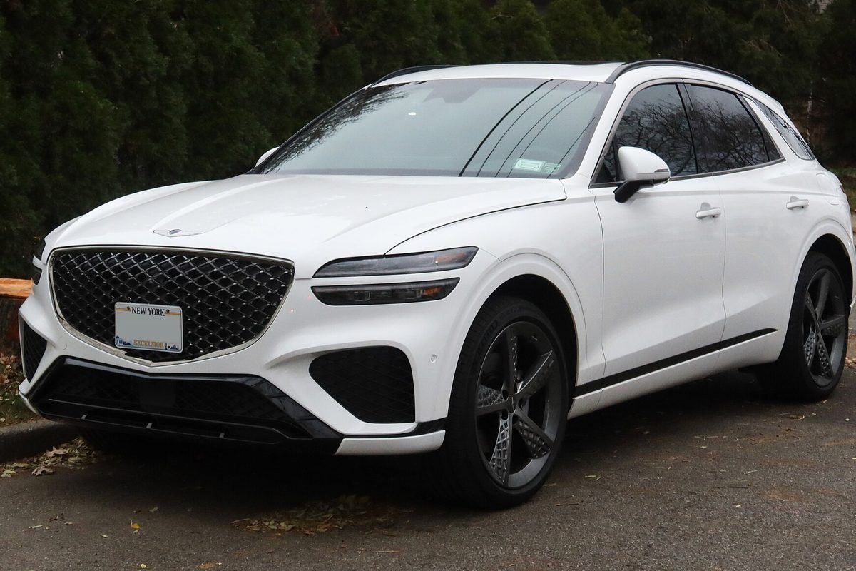 A white 2022 Genesis GV70 SUV with dark wheels, parked at a curb with fallen leaves and a tall hedge behind it