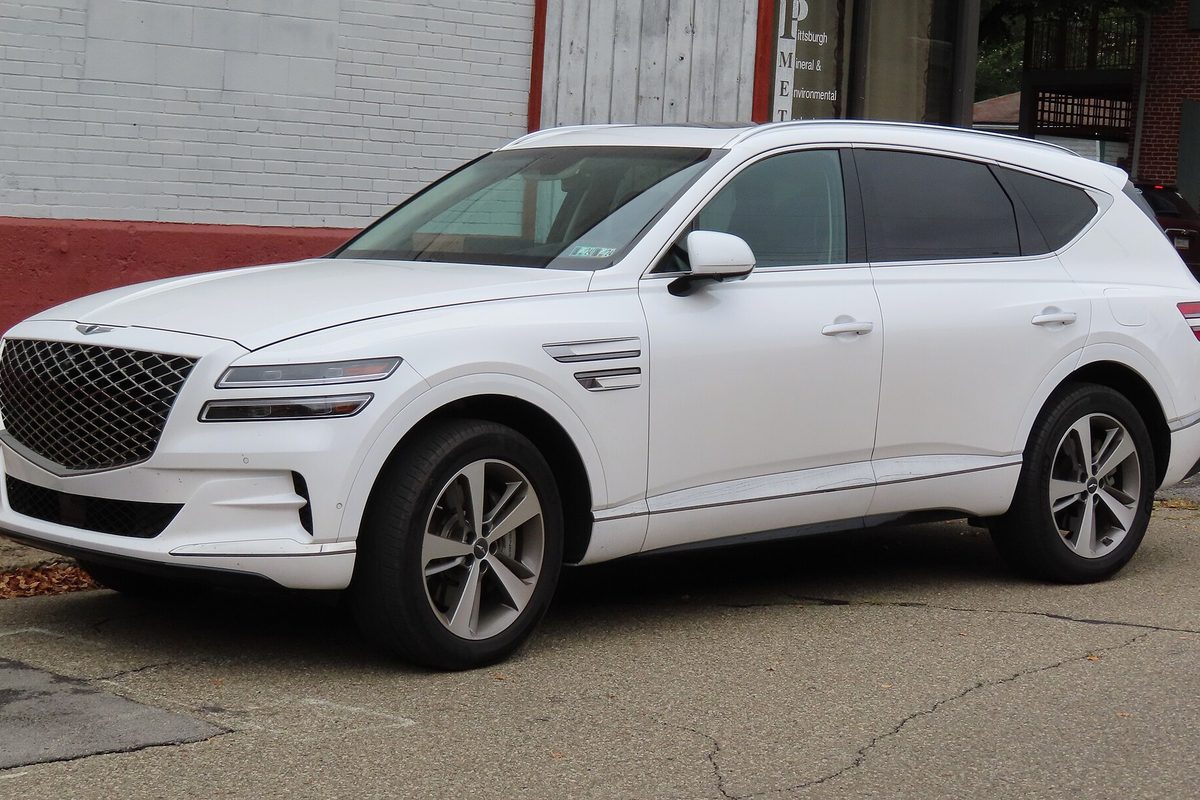 A white 2021 Genesis GV80 SUV parked at the curb beside a painted brick building