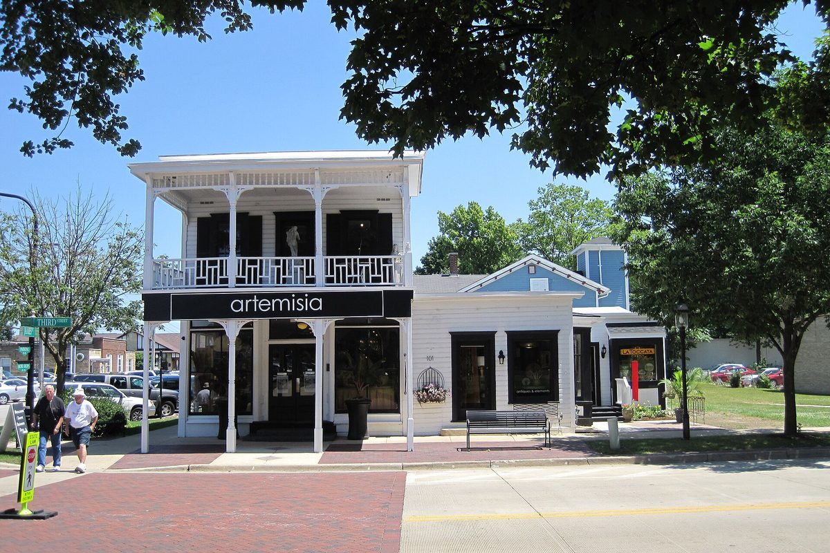 The Hull Building in Geneva's Central Geneva Historic District, a white two-story frame building with an upper balcony and a shop sign, on a brick sidewalk under shade trees