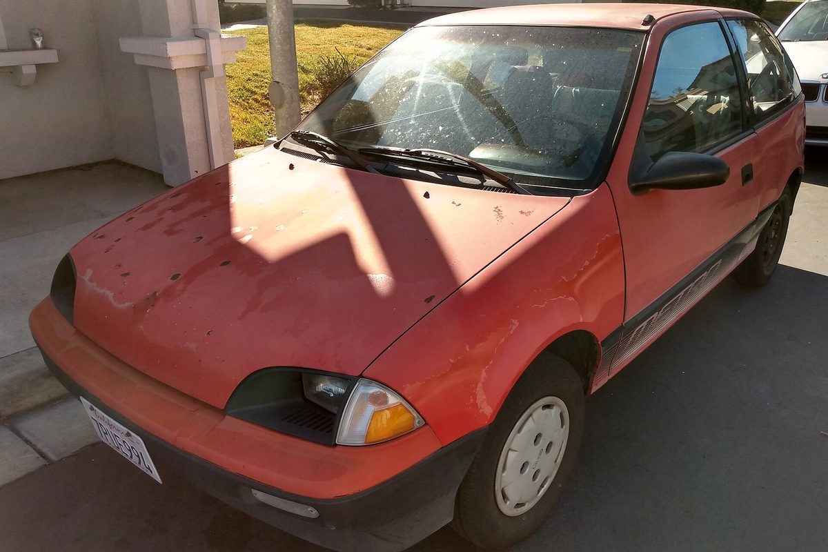 A faded red first-generation Geo Metro two-door hatchback with peeling paint on the hood, parked in a driveway