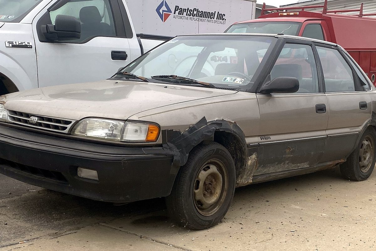 A faded tan first-generation Geo Prizm sedan with black tape over a rusted-out front fender and rust along the rocker panel
