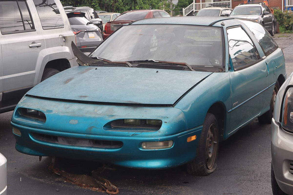 A faded turquoise Geo Storm wagon with its hood popped open and peeling clear coat, parked in a lot