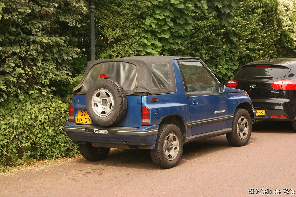 A blue Geo Tracker convertible with its black soft top up, seen from the rear with the spare tire mounted on the tailgate, parked on a brick driveway beside a tall hedge