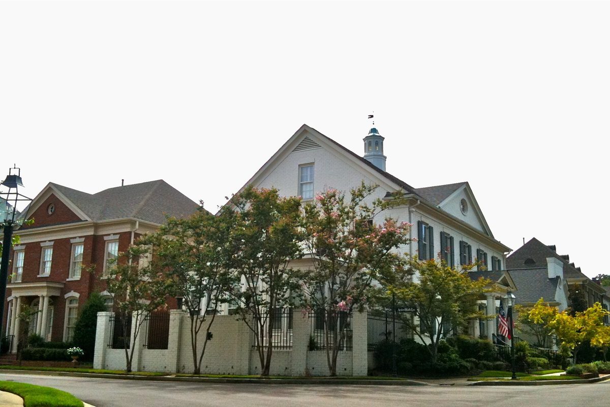 A white two-story house with a cupola and a red-brick house behind a low white wall and flowering crape myrtles on a curving Germantown street