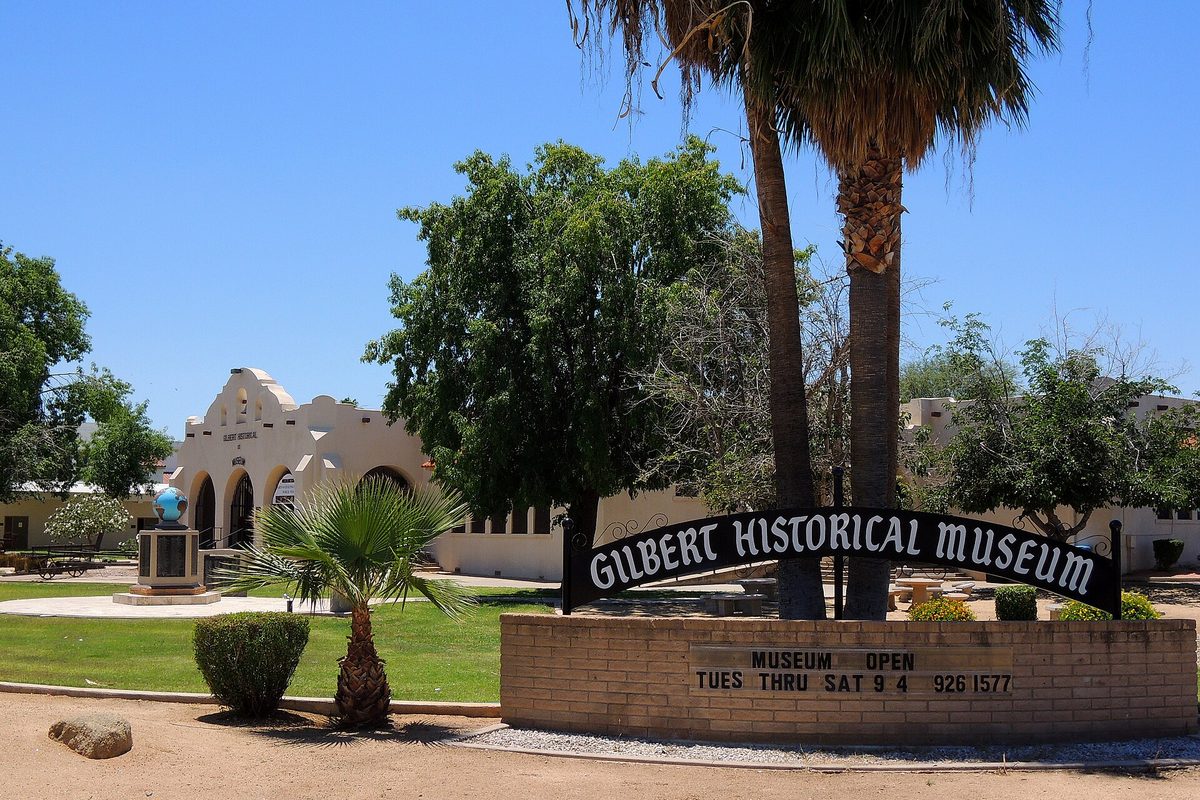 The white arched Gilbert Historical Museum behind palm trees and a lawn, with its sign arched over a brick base