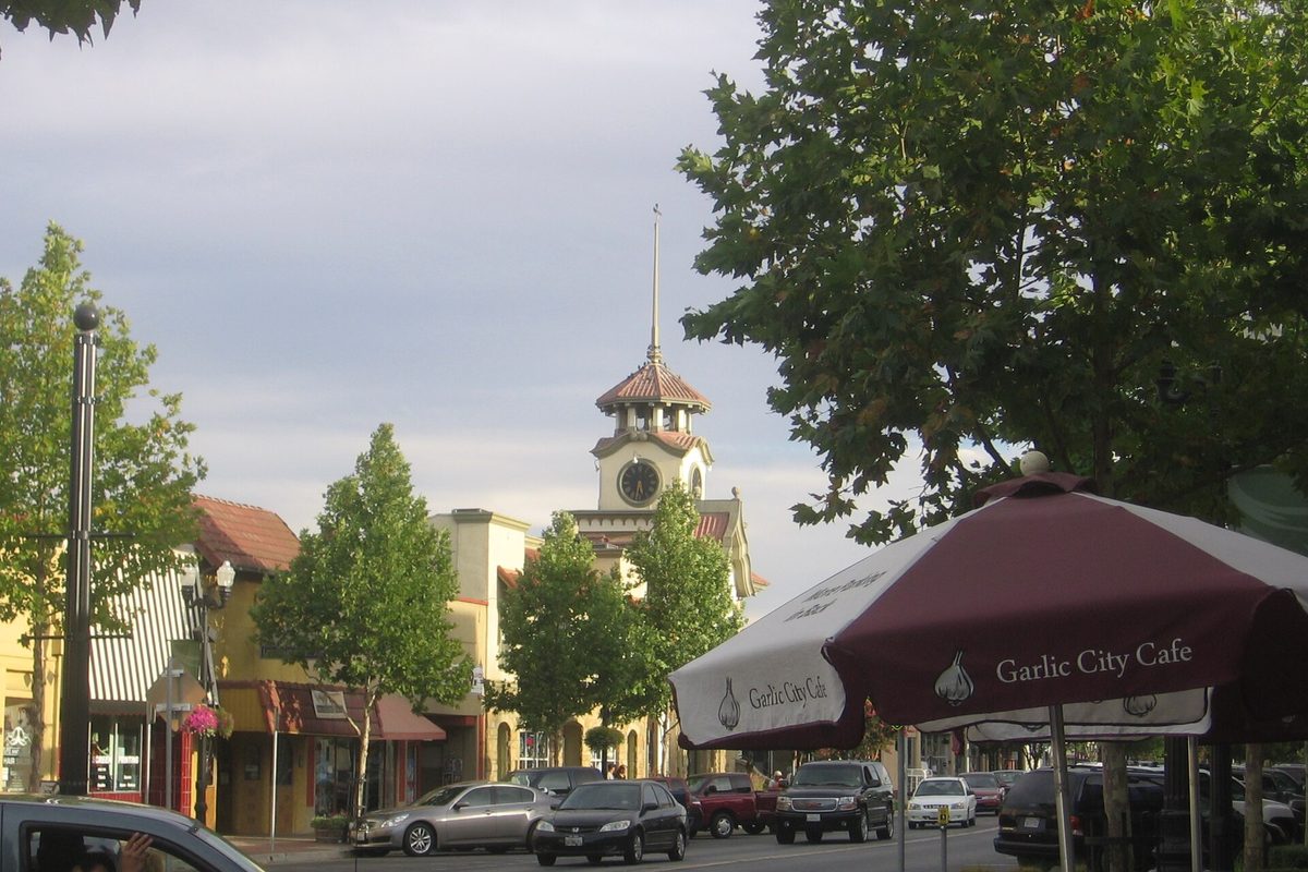 The clock tower of Gilroy's old 1905 City Hall rising above Monterey Street, with parked cars and a Garlic City Cafe umbrella in the foreground