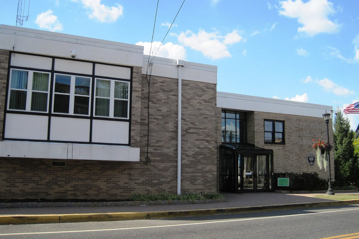 The Glassboro municipal building on South Main Street, a tan brick building with white panels, a glass entrance and an American flag, under a blue sky