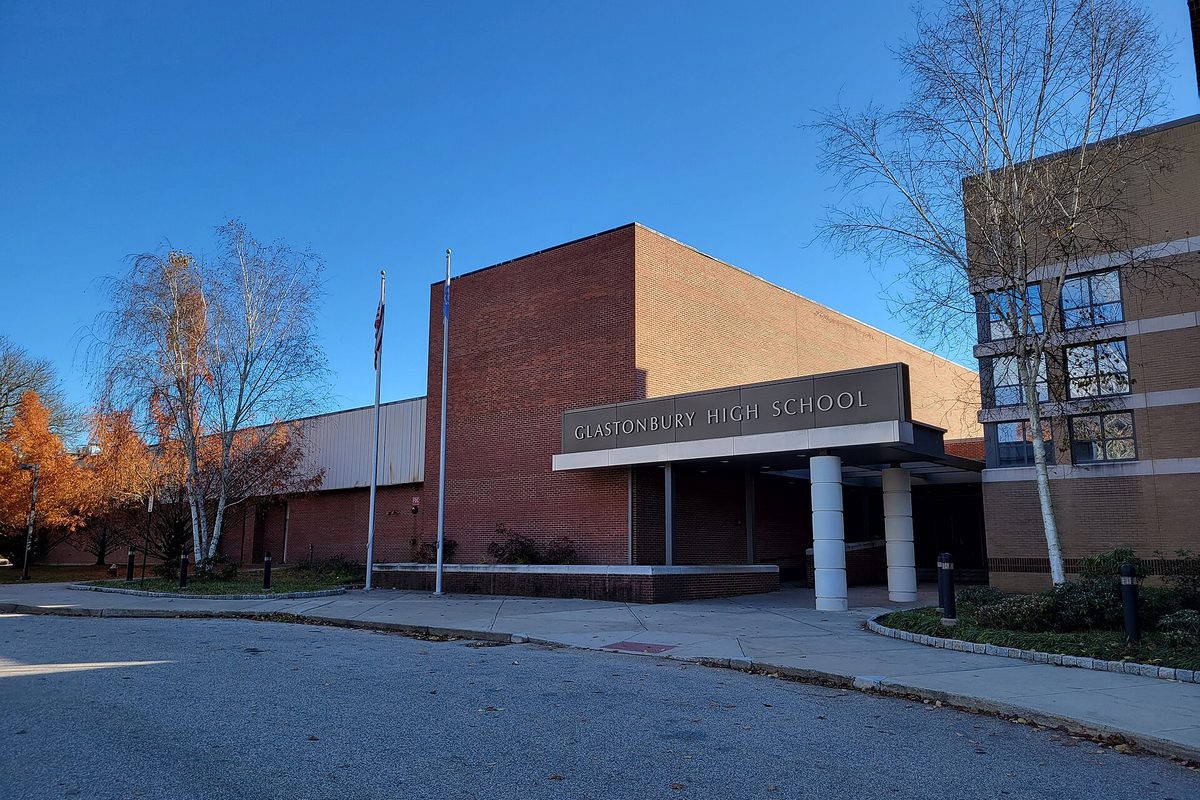 The brick front of Glastonbury High School with its entrance canopy and flagpoles, framed by autumn trees under a clear blue sky