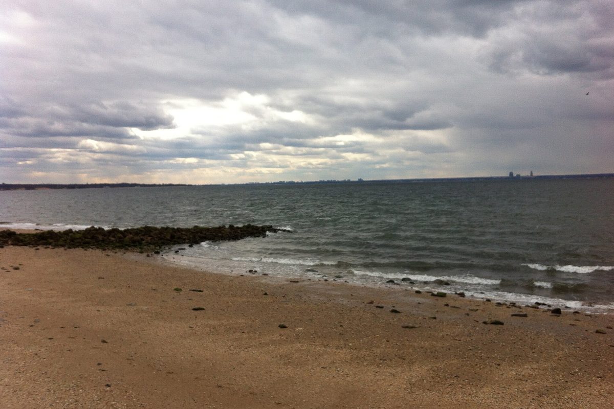 A sandy beach at Welwyn Preserve in Glen Cove, with a rock jetty running into gray water under heavy storm clouds