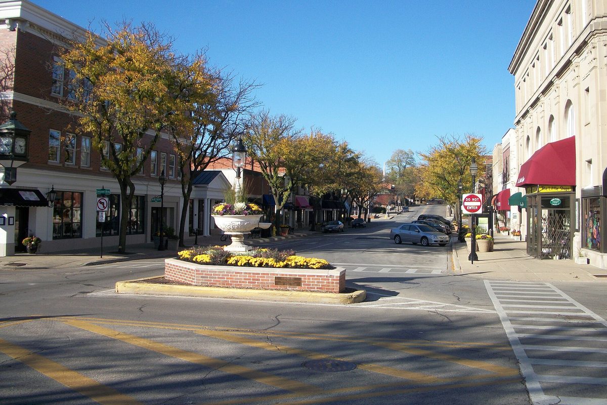 North Main Street in downtown Glen Ellyn on a sunny fall day, with a planter of yellow flowers in the road, brick storefronts and red awnings