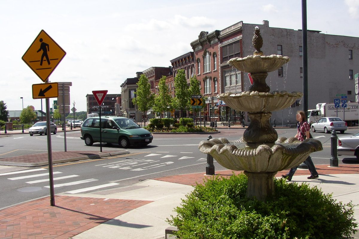Centennial Circle in downtown Glens Falls, with a tiered stone fountain in the foreground, a crosswalk sign, and brick storefronts along the roundabout