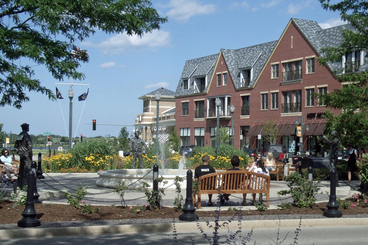 The plaza at The Glen Town Center in Glenview, with a fountain, bronze figures, flower beds and a bench in front of brick buildings with gray roofs