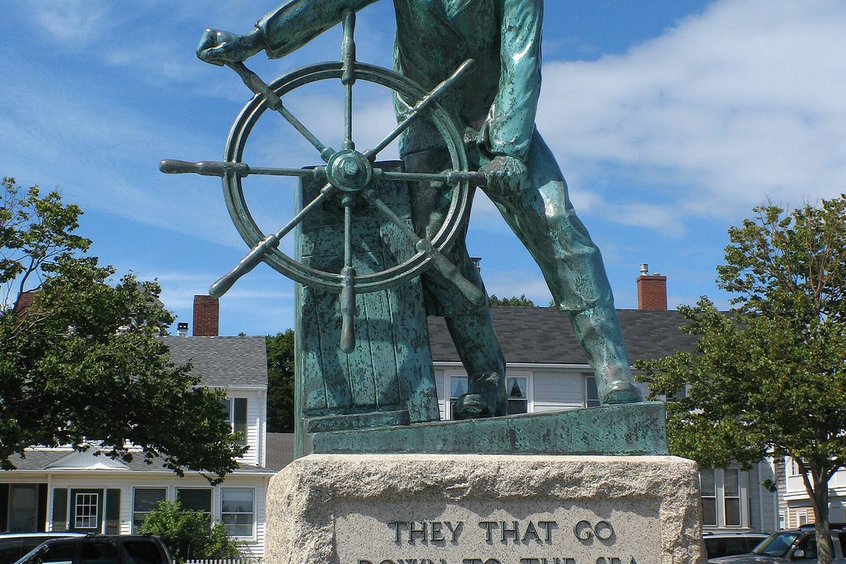 The bronze Fisherman's Memorial in Gloucester, a fisherman at a ship's wheel on a granite base carved They That Go Down to the Sea, with houses behind