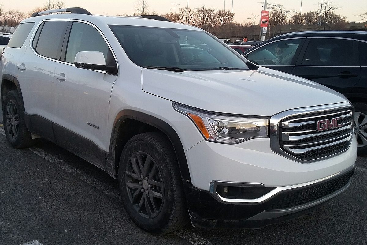 A white GMC Acadia crossover in a parking lot at dusk, a black SUV behind it