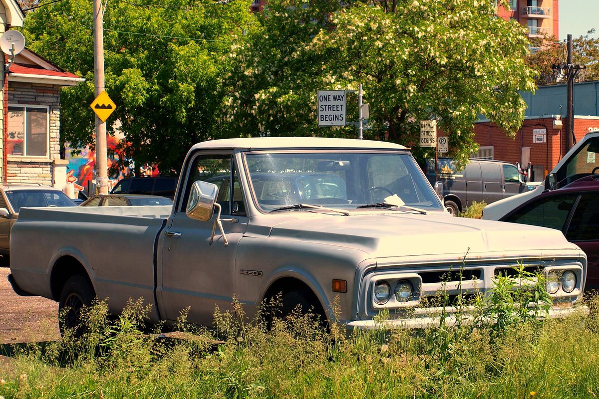 A faded silver older-generation GMC C/K regular-cab pickup parked in tall weeds beside a street