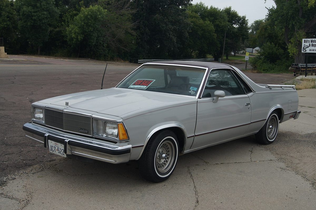 A silver GMC Caballero car-based pickup with a dark roof, wire wheels and whitewall tires, parked on a cracked asphalt lot by trees