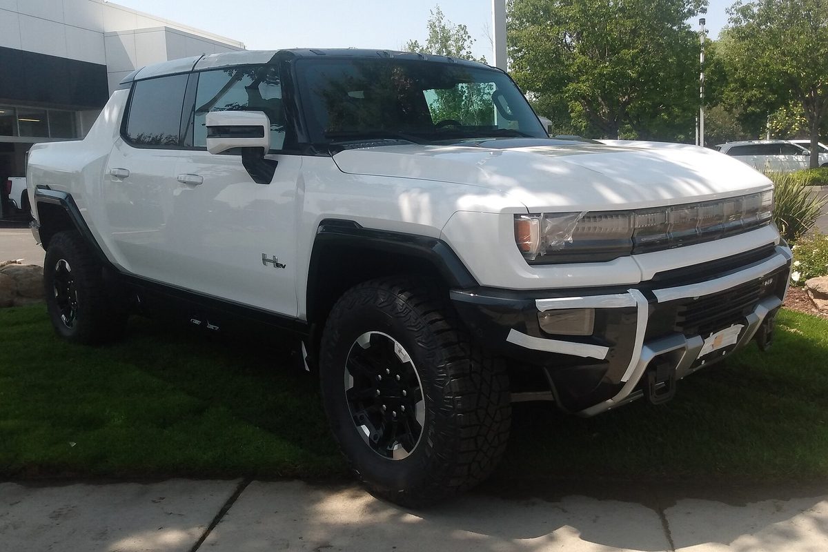 A white GMC Hummer EV pickup with black fender flares and off-road tires, parked on grass outside a dealership