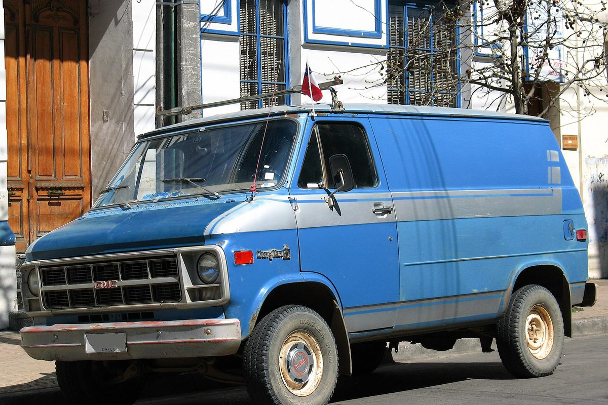 A blue GMC van with a silver side band and round headlights, parked on a street in front of an old building with tall wooden doors