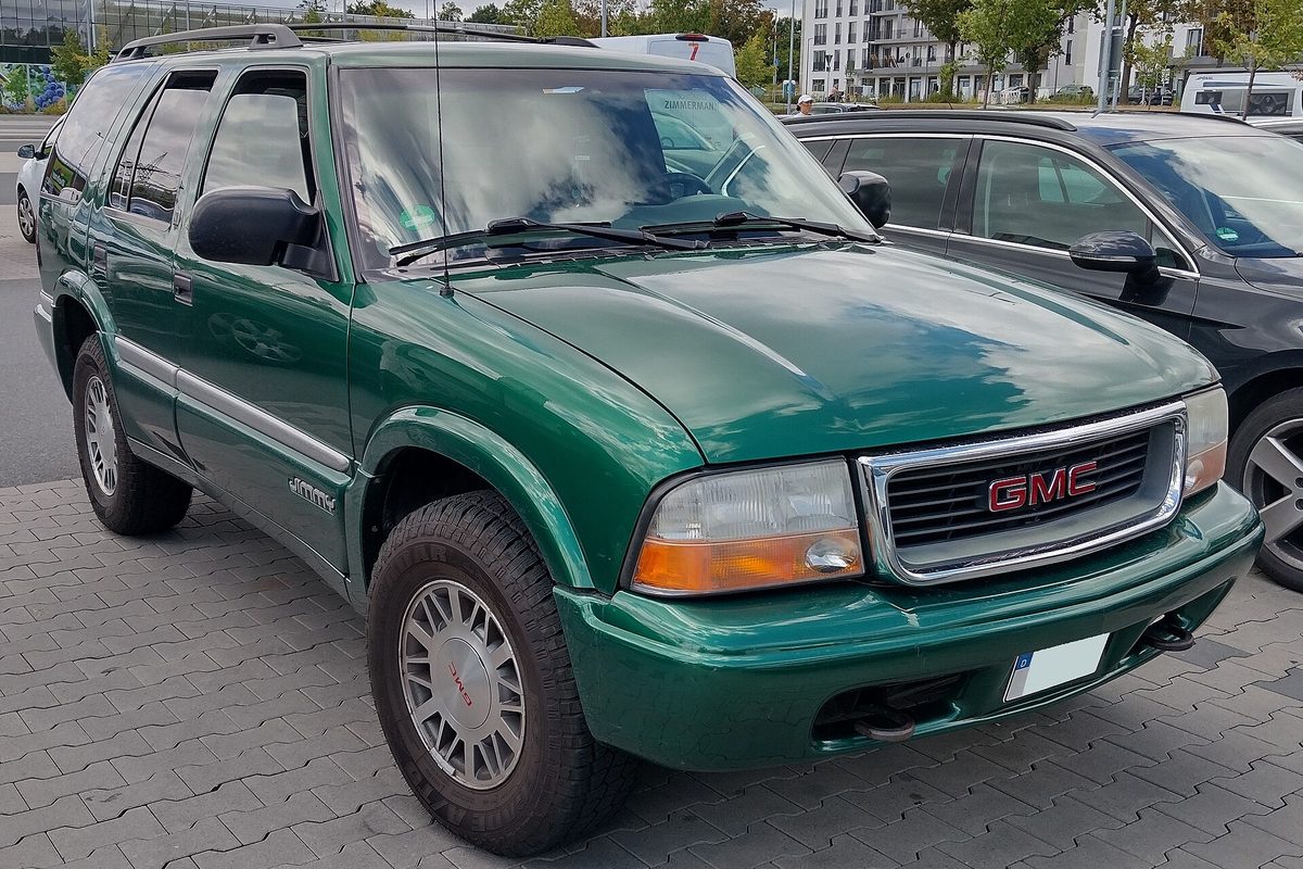 A green four-door GMC Jimmy, the SUV from the S-series family, parked on a paved lot beside other cars