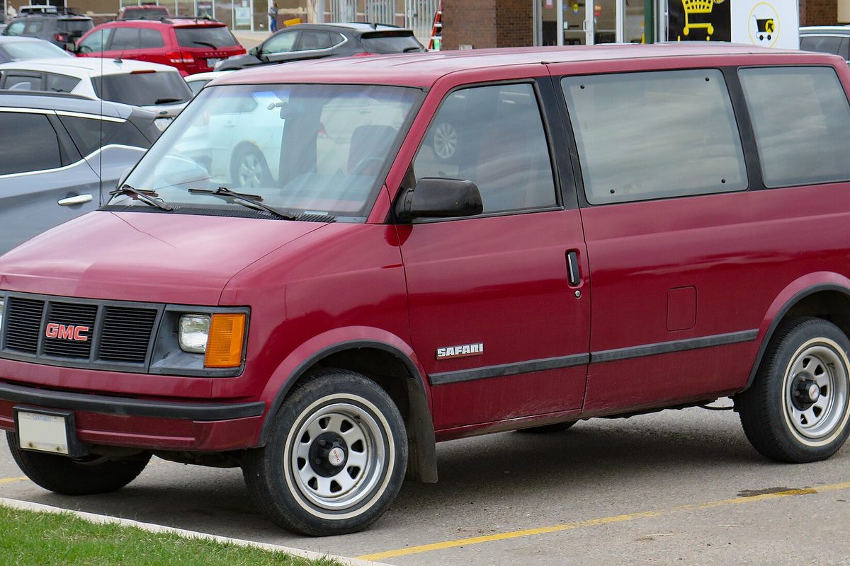 A deep red first-generation GMC Safari van with steel wheels, parked in a store lot