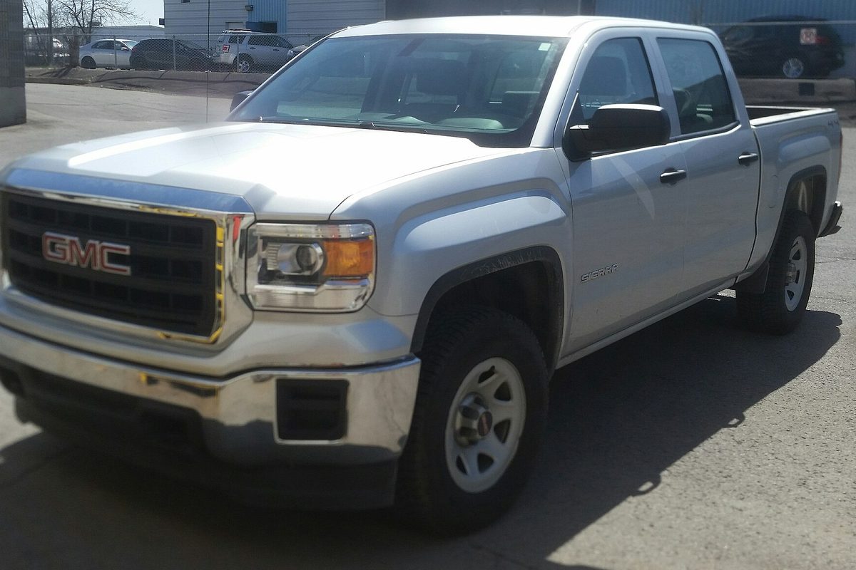 A silver four-door GMC Sierra 1500 pickup on plain steel wheels, parked on a gray lot with other cars behind it