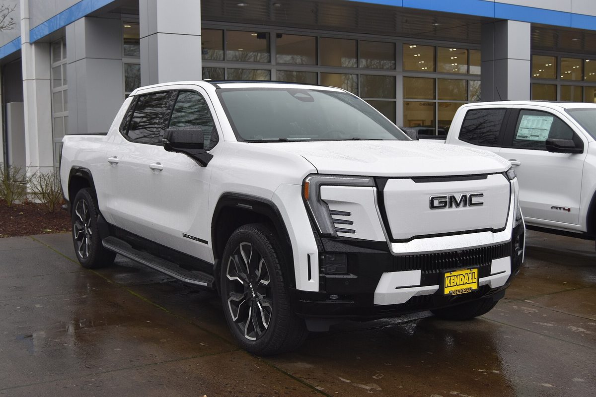 A white GMC Sierra EV crew-cab pickup with a white panel grille and running boards, parked on wet pavement outside a dealership