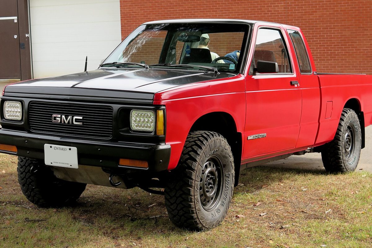 A red and black GMC Sonoma extended-cab pickup on large off-road tires, parked on grass by a brick building