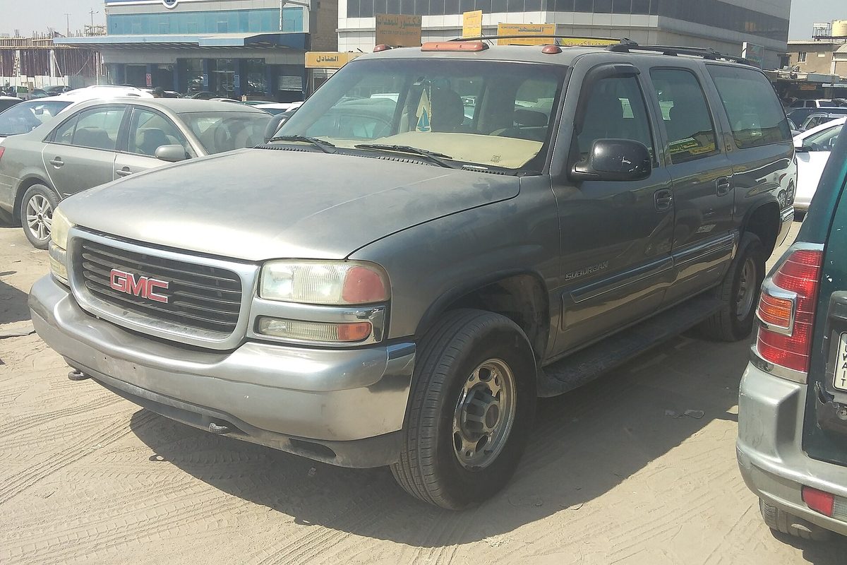 A light pewter GMC Suburban with a dusty hood and faded paint, parked on sand in a crowded lot