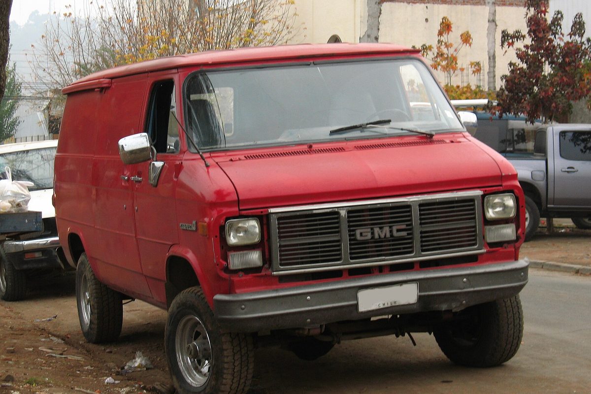 A red GMC Vandura cargo van with no side windows, a chrome GMC grille and rectangular headlights, parked at a curb