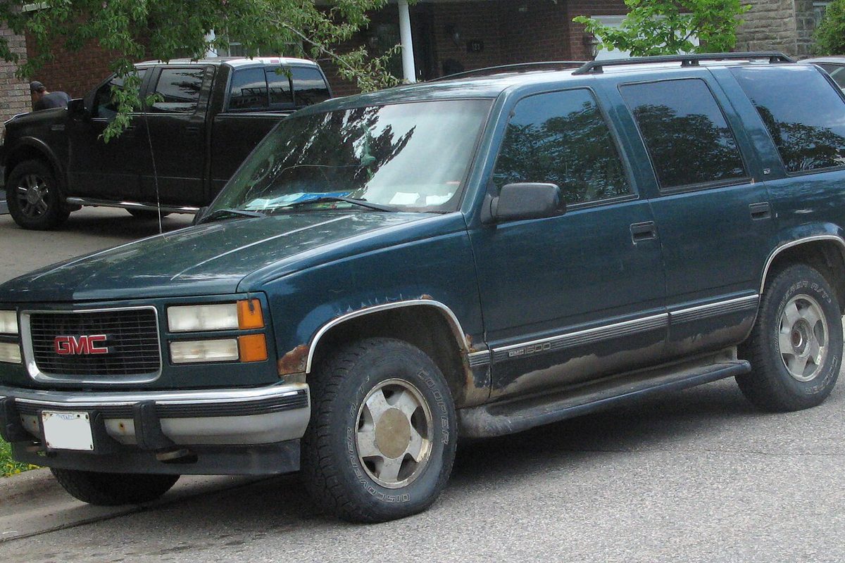 A dark green late-1990s GMC Yukon five-door with rust along the lower doors, parked at a curb