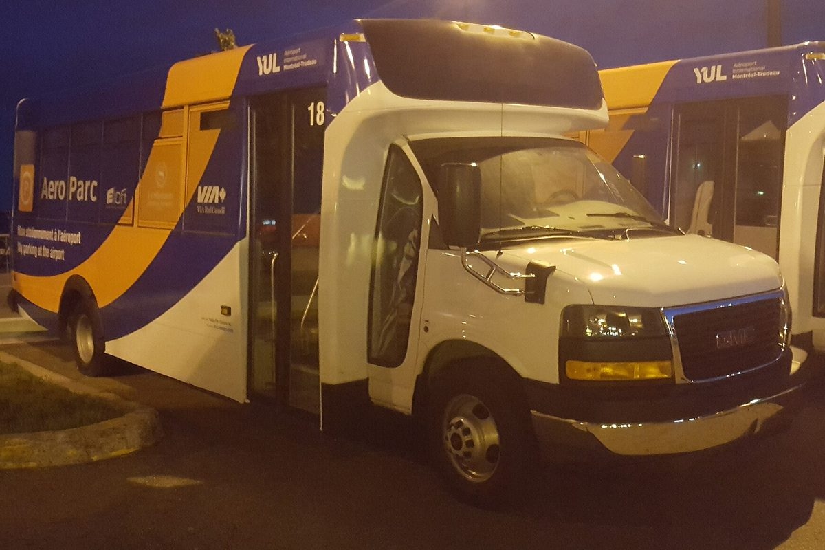 A white GMC Savana cutaway shuttle bus with a blue and orange airport parking livery, parked at night in Dorval, Quebec
