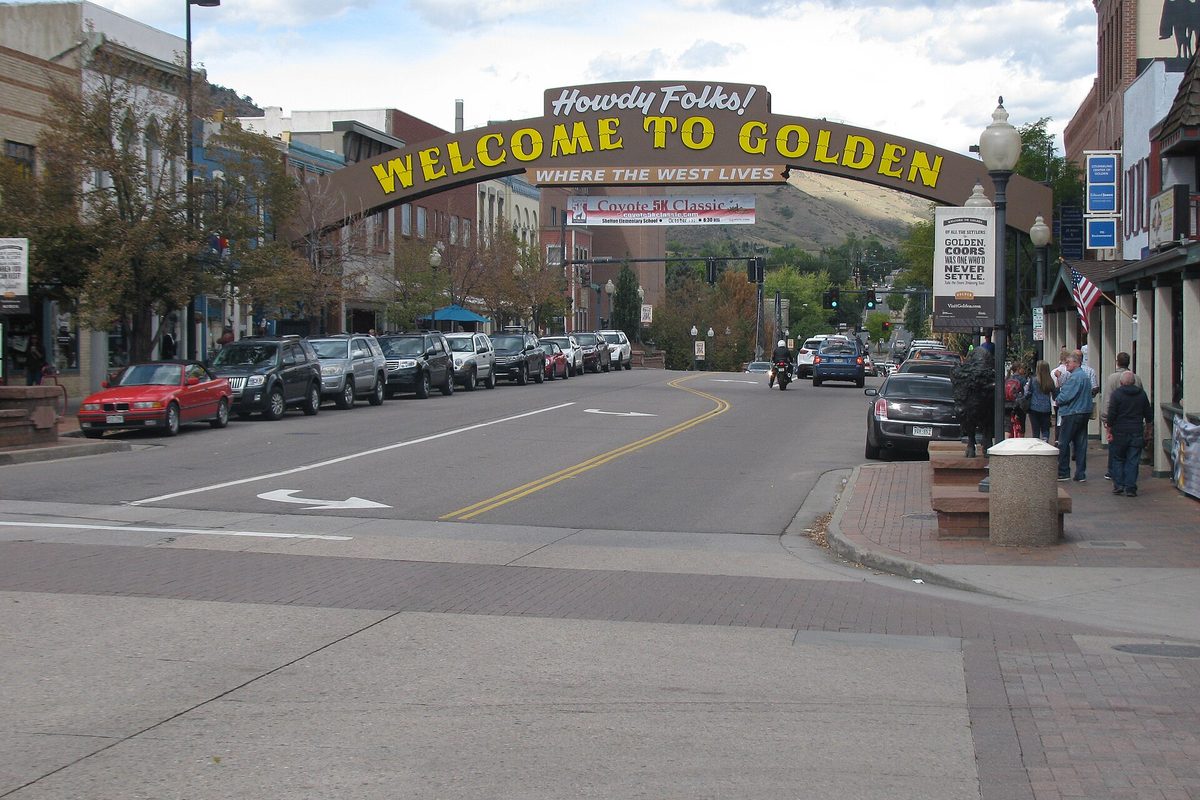 The Howdy Folks! Welcome to Golden arch over Washington Street, with cars parked along the curb and a foothill in the background