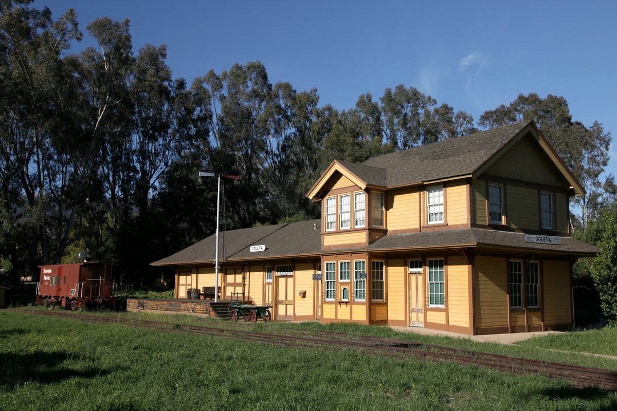 The yellow two-story wooden Goleta train depot with a red caboose beside it, a green lawn in front and tall eucalyptus trees behind