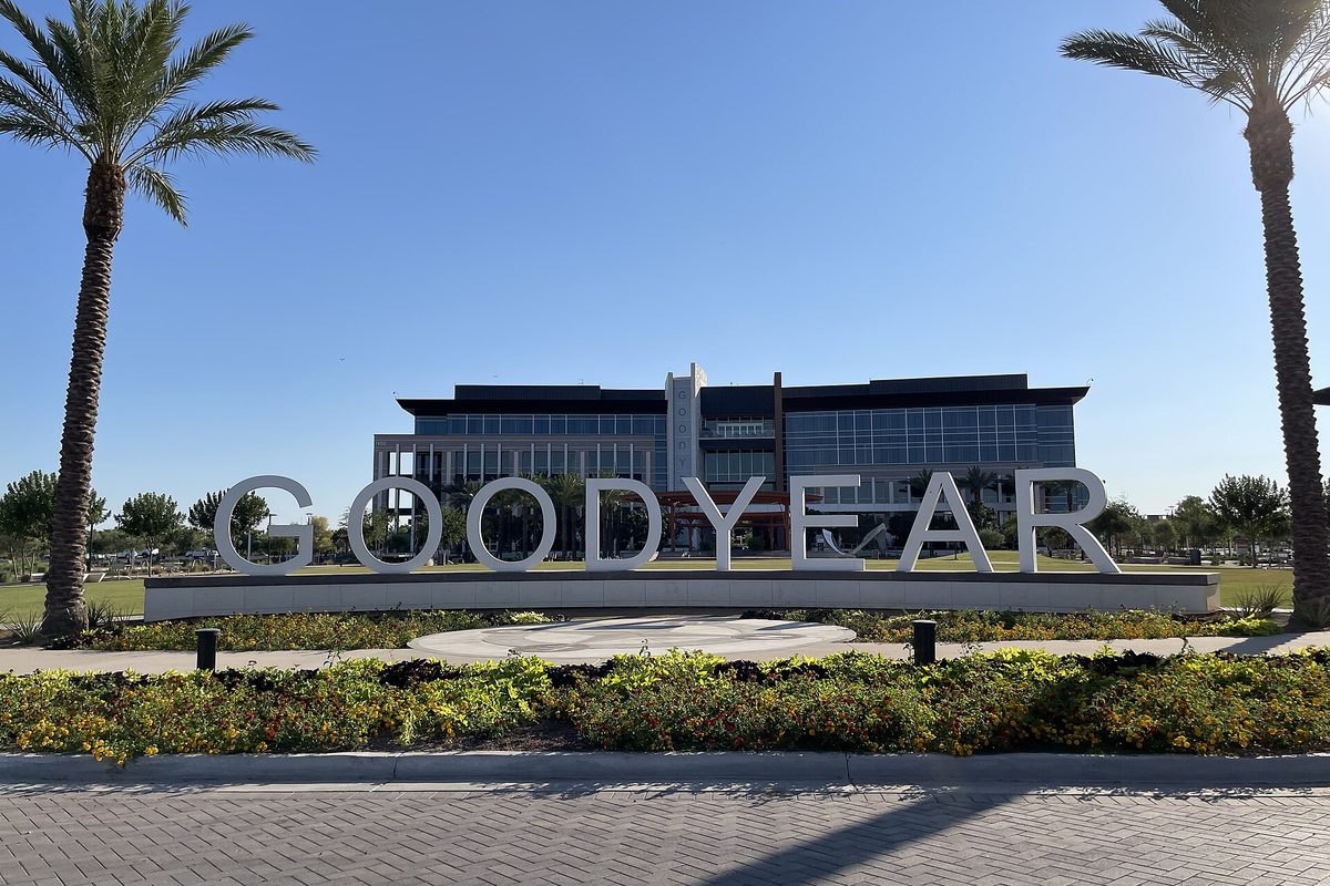Large white GOODYEAR letters in front of Goodyear's glass-fronted city hall, framed by palm trees and a bed of yellow flowers