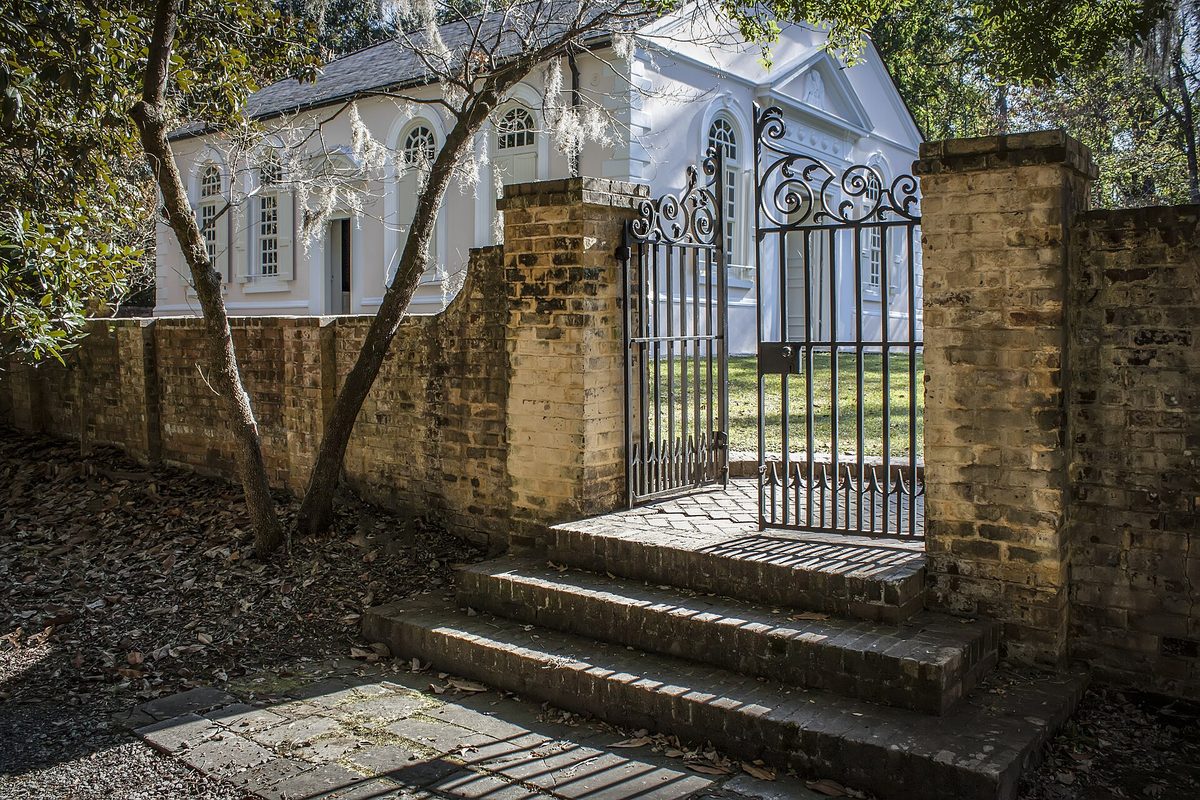 An old brick wall and open iron gate in front of the white St. James Goose Creek Parish Church, with moss hanging from the trees