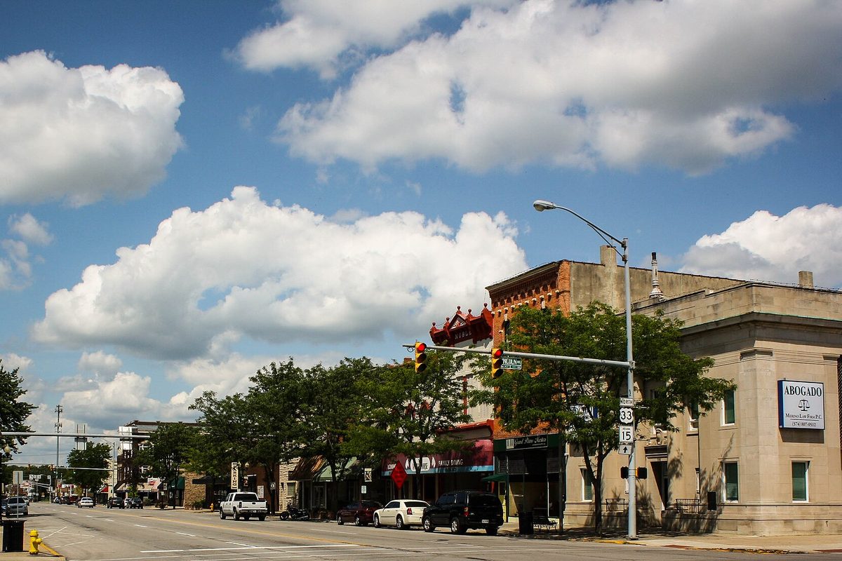 Main Street at Lincoln Avenue in downtown Goshen, with brick storefronts, trees, a U.S. 33 sign on a signal pole and white clouds in a blue sky