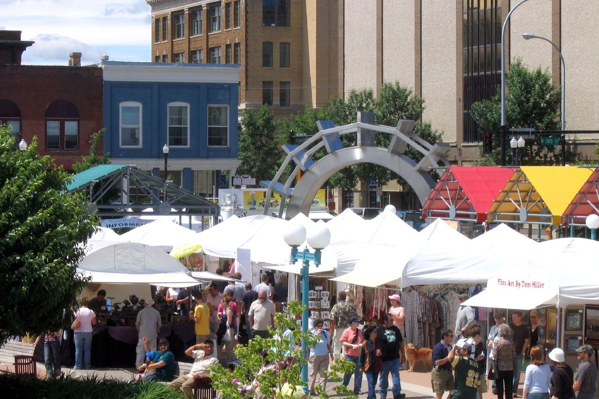 Crowds walk between white vendor tents at the Grand Cities Art Fest in downtown Grand Forks, with a steel arch sculpture and brick buildings behind