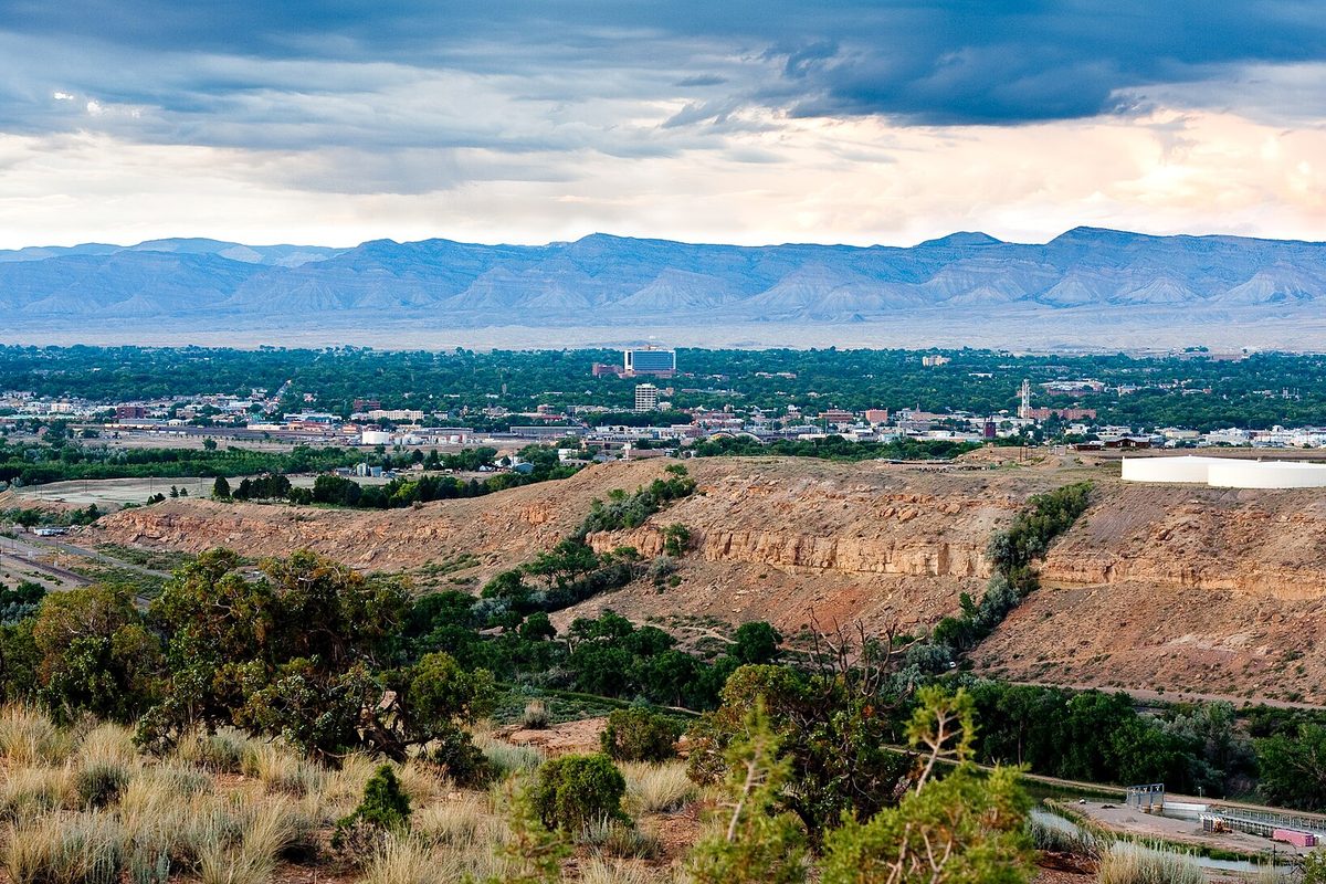 Grand Junction spread across a green valley, seen from a dry, grassy bluff, with long flat-topped mountains under dark storm clouds behind the city