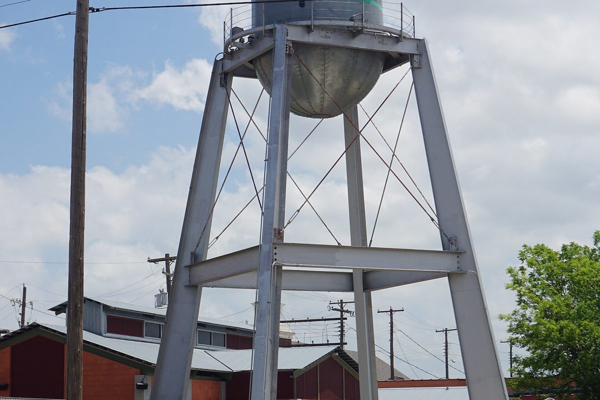 The silver steel Market Square water tower in Grand Prairie on braced legs above red-walled buildings and power lines