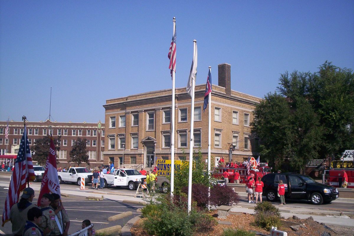 Granite City City Hall, a tan brick building, with three flagpoles out front, a parade crowd in red shirts, pickup trucks and a fire engine under a clear sky