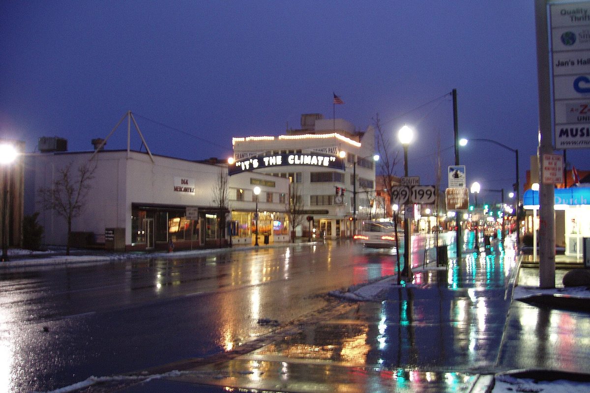 The lit It's the Climate sign over a downtown Grants Pass street at dusk, with wet pavement, streetlights and patches of snow