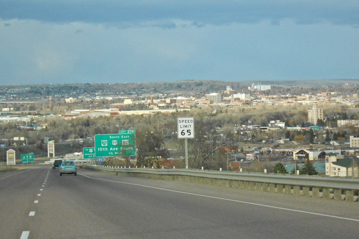 Interstate 15 heading into Great Falls past a speed limit 65 sign and a green exit sign for 10th Ave South, with the city spread across the valley beyond
