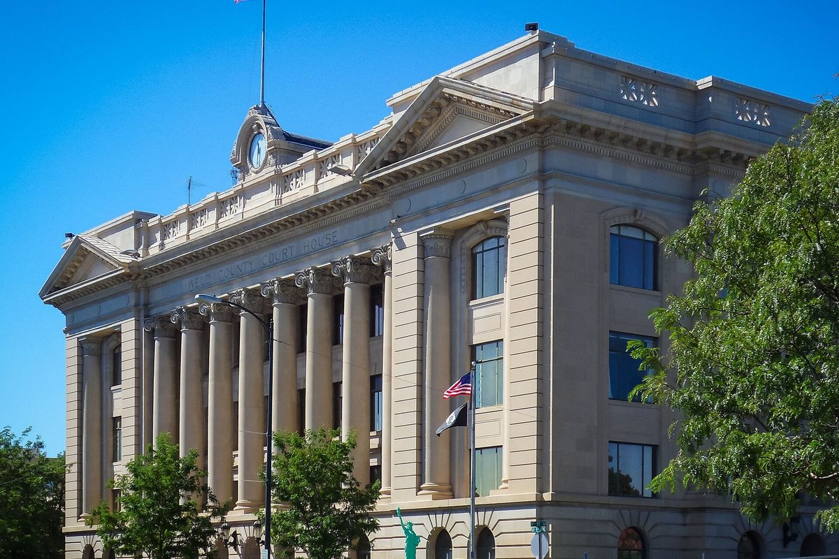 The columned stone courthouse in Greeley with a clock above the roofline, an American flag and leafy trees in front, under a clear blue sky