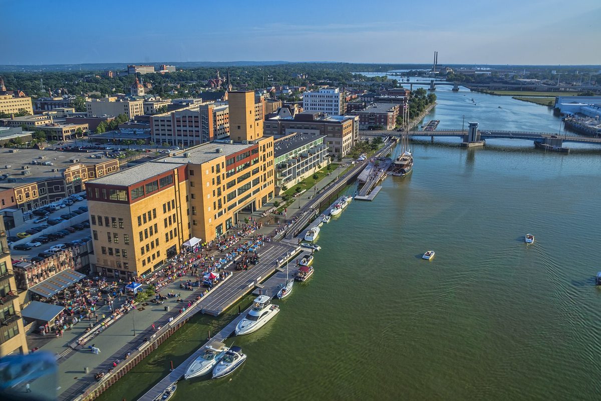 Aerial view of downtown Green Bay and a crowd on the CityDeck along the Fox River, with boats docked and a bridge in the distance