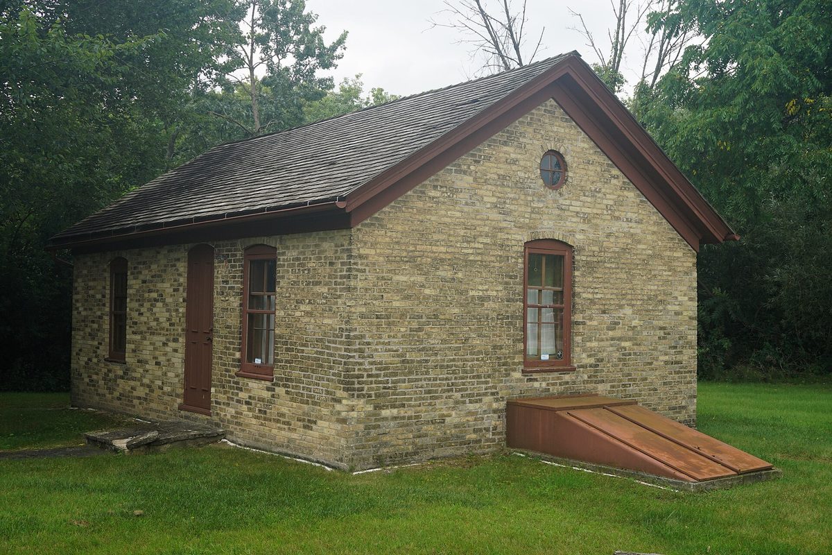A small cream brick building with a red door, arched windows and a round attic window, home of the Greenfield Historical Society, among tall trees