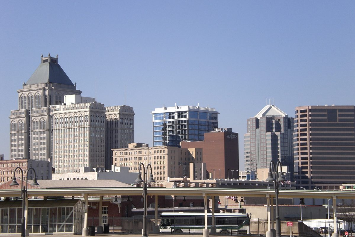 Downtown Greensboro skyline on a clear day, older stone towers beside glass office buildings, seen past a covered platform and street lamps