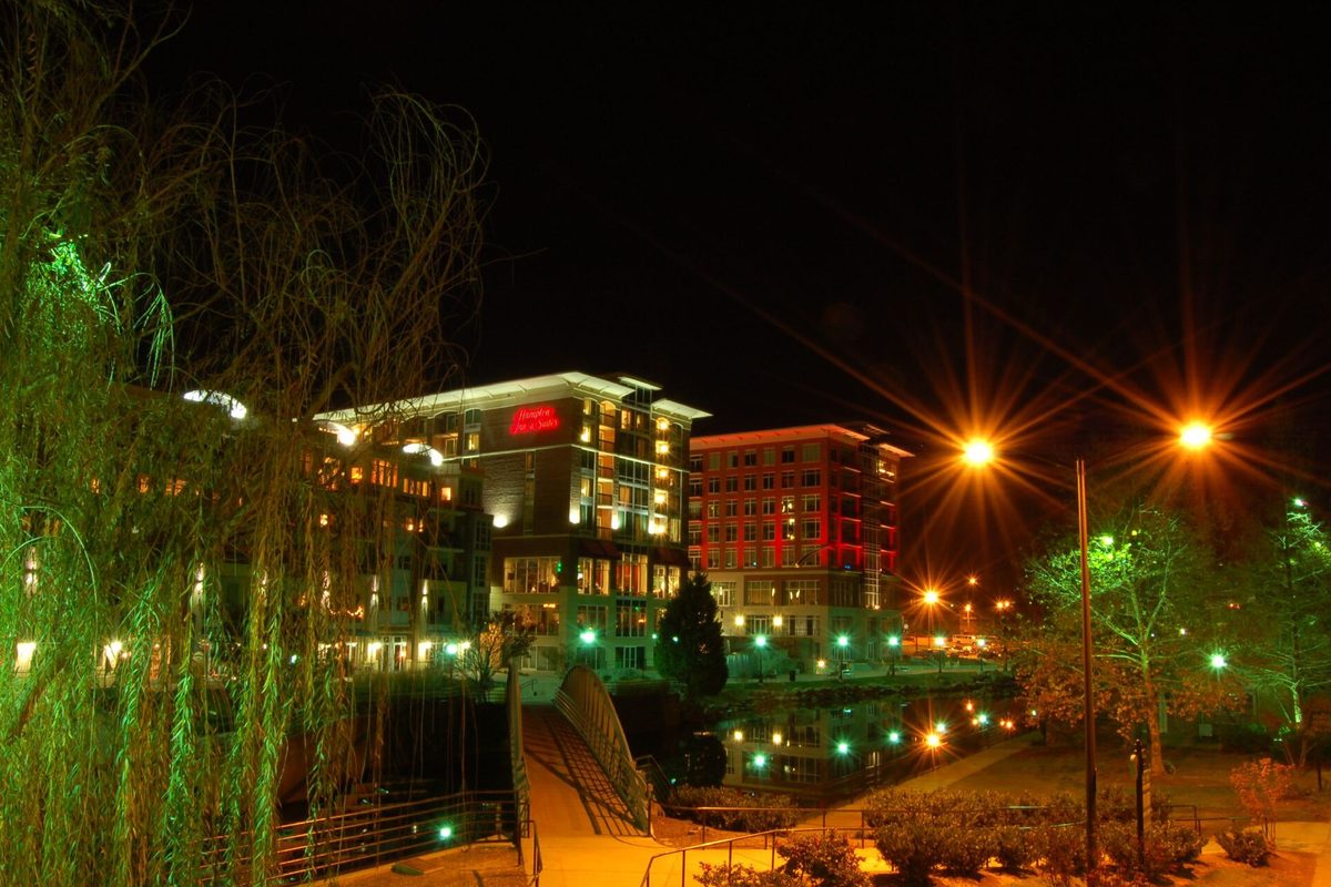 RiverPlace buildings in downtown Greenville lit up at night beside the Reedy River, with a footbridge, willow branches and glowing street lamps