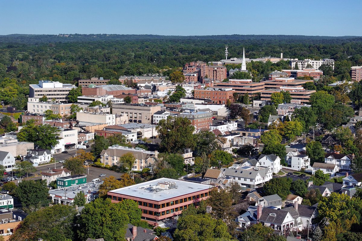 Aerial view of downtown Greenwich: brick office buildings and a white church steeple among thick green trees, with houses in the foreground